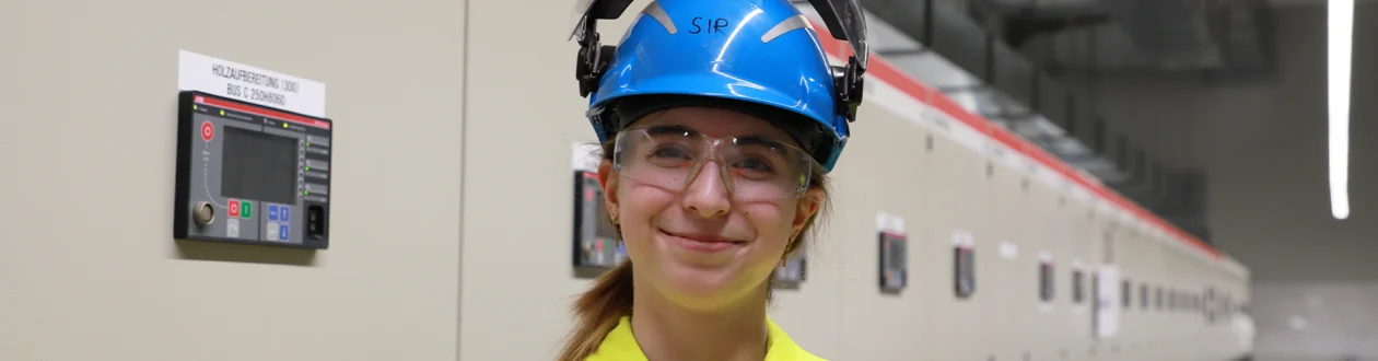 Smiling young woman in yellow PPE, blue helmet and safety goggles in an industrial plant with control cabinets.