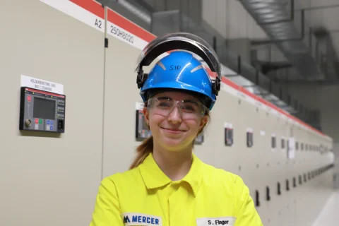 Smiling young woman in yellow PPE, blue helmet and safety goggles in an industrial plant with control cabinets.