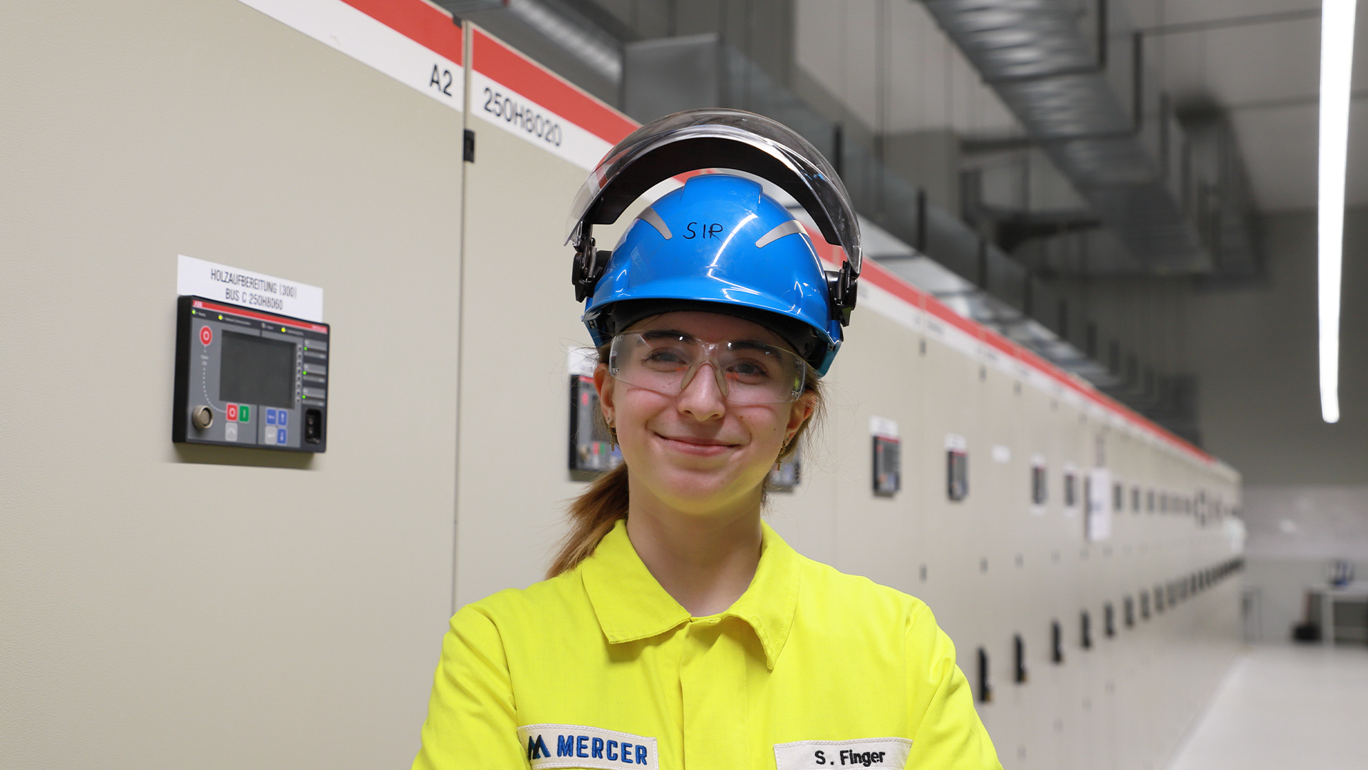 Smiling young woman in yellow PPE, blue helmet and safety goggles in an industrial plant with control cabinets.