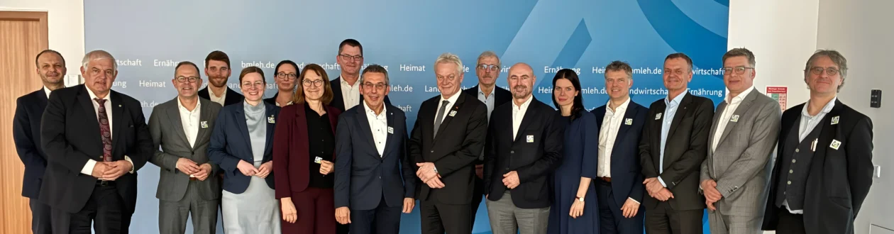 Large group of smiling people, mostly men in business attire, many with lapel pins, in front of a blue wall with German text.