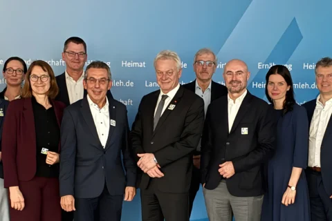 Large group of smiling people, mostly men in business attire, many with lapel pins, in front of a blue wall with German text.