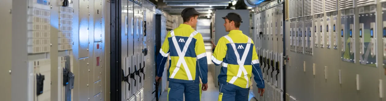 Two technicians in high-vis jackets and caps walk through a server room lined with grey cabinets. One gestures while talking.