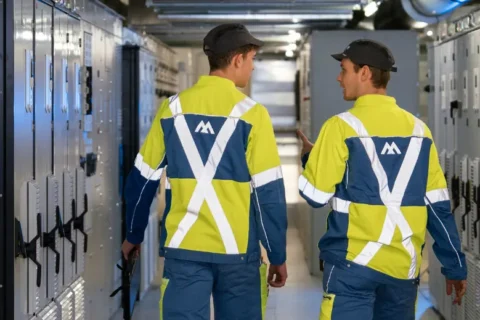 Two technicians in high-vis jackets and caps walk through a server room lined with grey cabinets. One gestures while talking.