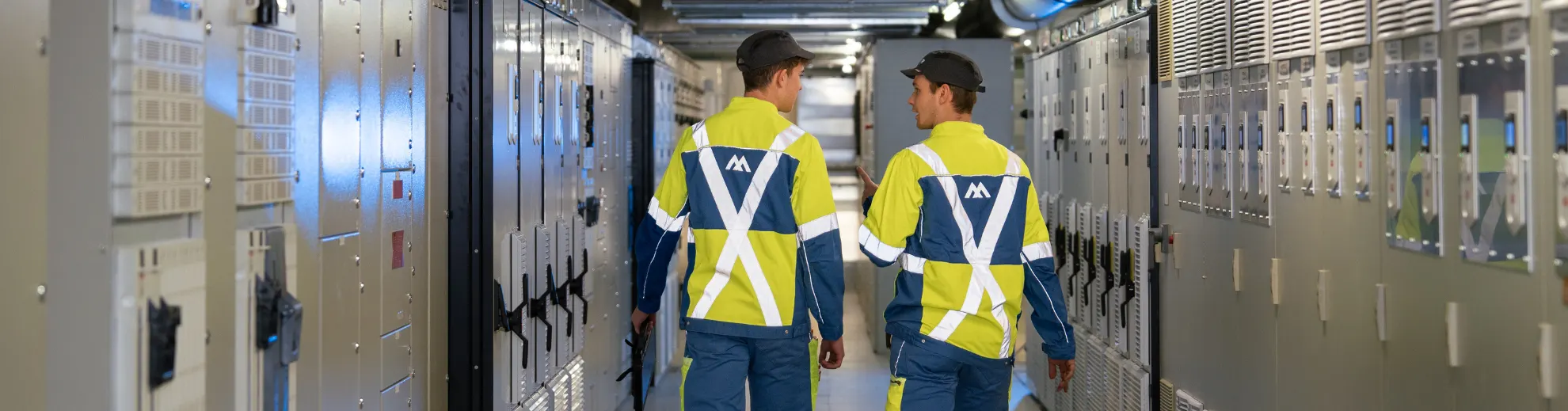 Two technicians in high-vis jackets and caps walk through a server room lined with grey cabinets. One gestures while talking.