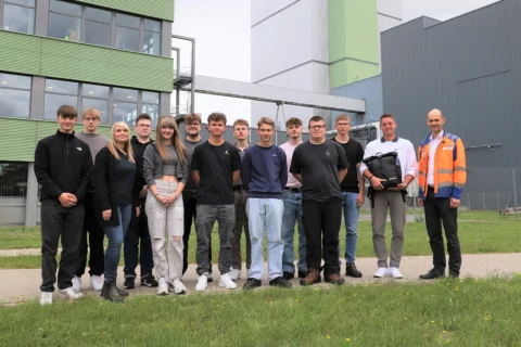Smiling group of 11 students and 2 instructors (one in safety vest) outdoors by green/gray industrial buildings.