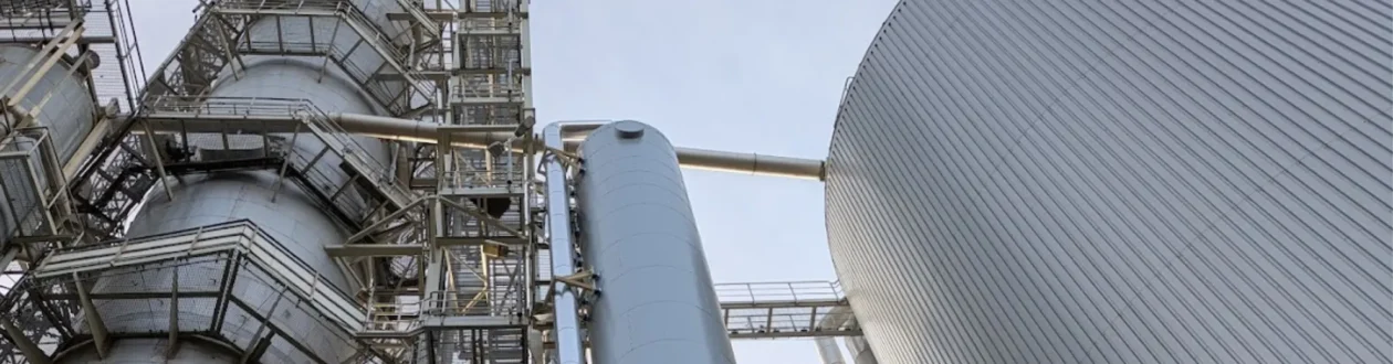 Large industrial facility with a multi-level processing tower, a tall silver tank, and a massive corrugated storage tank under a pale sky.