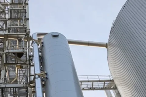 Large industrial facility with a multi-level processing tower, a tall silver tank, and a massive corrugated storage tank under a pale sky.