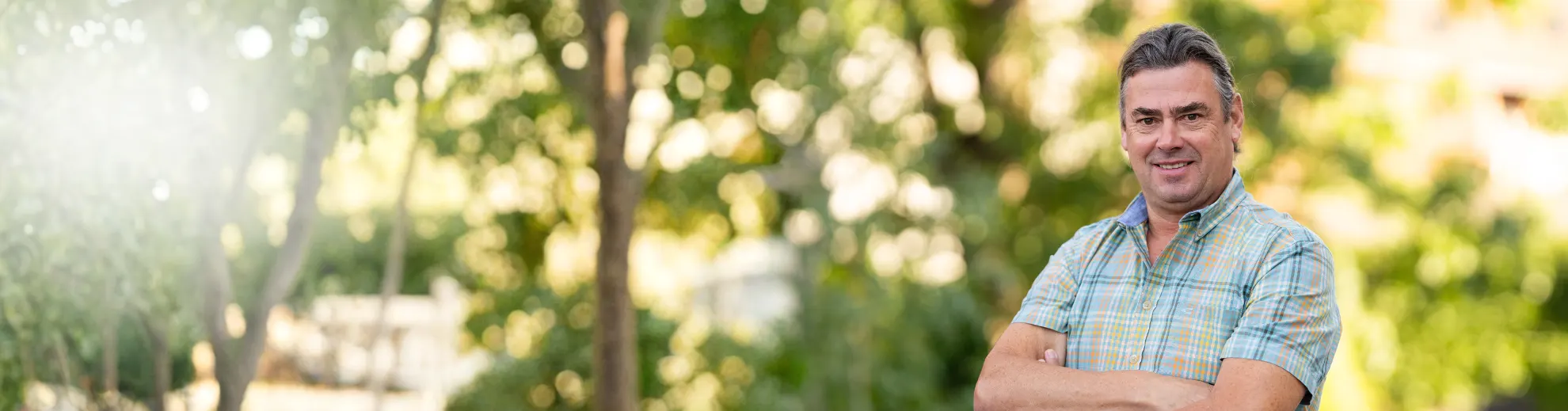 A smiling middle-aged man in a plaid shirt with crossed arms stands against a blurred green outdoor background with sun flare.