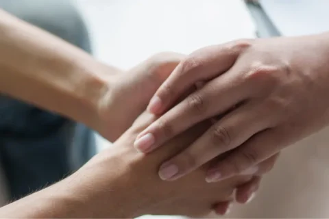 Close-up: A hand in a blue shirt comfortingly holds the hand of a person in dark clothing. Symbolizes support.