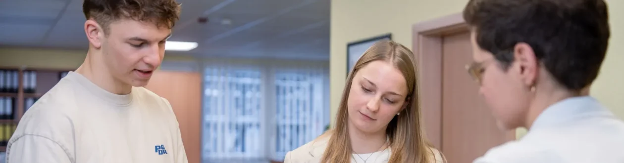 Three people. A young man and a woman are looking intently at something. Another person wearing glasses is looking at the woman. Office environment.